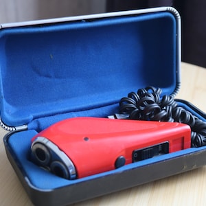 Könnte beinhalten: Ein roter elektrischer Rasierer mit einem schwarzen Kabel und einem schwarzen Etui mit blauem Innenfutter.