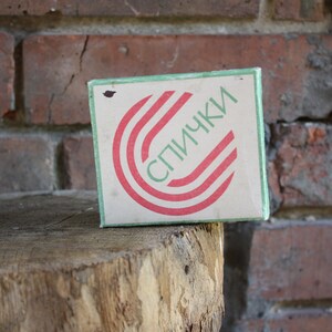 Pode incluir: Uma caixa de fósforos com um design vermelho e verde. A caixa diz "СПИЧКИ" em letras cirílicas.
