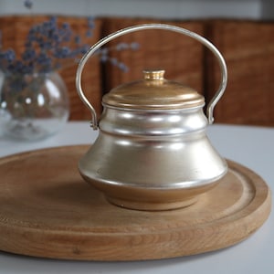 Puede incluir: Una pequeña olla de metal plateado con tapa y asa doradas. La olla está sobre una tabla de cortar redonda de madera. El fondo incluye un jarrón con lavanda y una cesta de mimbre marrón.