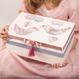 Può includere: Una scatola rettangolare bianca con coperchio rosa e bordo decorativo. Il coperchio presenta due uccelli illustrati con cuori e nuvole. Un nastro e un nappa rosa adornano la parte anteriore. La scatola è tenuta da una persona che indossa un vestito rosa.