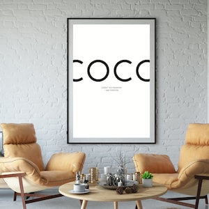 Peut inclure: Une affiche encadrée en noir et blanc avec le mot "COCO" en grandes lettres grasses. En dessous, le texte "I DON'T DO FASHION I AM FASHION". L'affiche est accrochée à un mur de briques blanches.