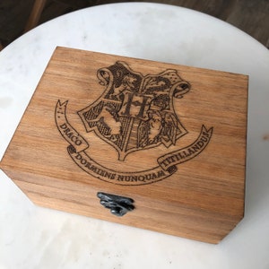 May include: A wooden box with a hinged lid and a metal latch. The top of the box is decorated with a laser-engraved Hogwarts crest and the motto "Draco Dormiens Nunquam Titillandus".