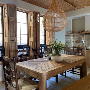 May include: A wooden dining table with a chevron pattern top and six chairs. The table is set with a bowl of fruit and a bottle of wine. The room has a rustic feel with exposed wooden beams and large windows.