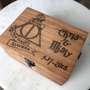 May include: A wooden box with a dark finish, featuring engraved text and symbols. The box has a clasp and is decorated with the words "Chris & Misty" and the date "9.17.2018". The top of the box also has the Deathly Hallows symbol and the words "All this time? Always."