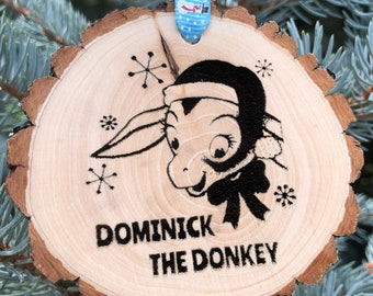 May include: A wooden Christmas ornament with a black and white illustration of a donkey wearing a Santa hat. The ornament is decorated with snowflakes and the text "Dominick the Donkey".