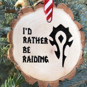 Könnte beinhalten: Ein hölzerner Weihnachtsbaumschmuck mit einem rot-weiß gestreiften Band. Der Schmuck ist eine runde Holzscheibe mit dem Text "I'd rather be raiding" und einem schwarzen Symbol.
