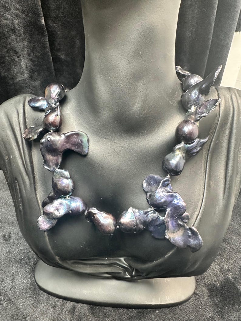 May include: A necklace made of iridescent black pearls. The pearls are strung together in a loose, flowing style. The necklace is displayed on a black mannequin bust.