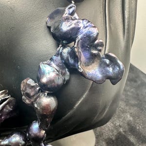 May include: A necklace made of large, irregular, iridescent black pearls. The pearls are strung together with a silver or white metal thread. The necklace is displayed on a black mannequin bust.