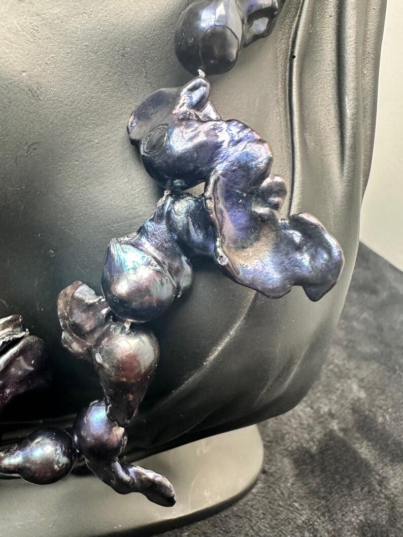 May include: A necklace made of large, irregular, iridescent black pearls. The pearls are strung together with a silver or white metal thread. The necklace is displayed on a black mannequin bust.