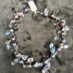 May include: A necklace made of dark iridescent baroque pearls. The pearls are strung on a silver chain and are of varying sizes and shapes. The necklace is displayed on a dark brown surface. The tag attached to the necklace reads "Real Cultured South Sea Pearls 9-10mm".
