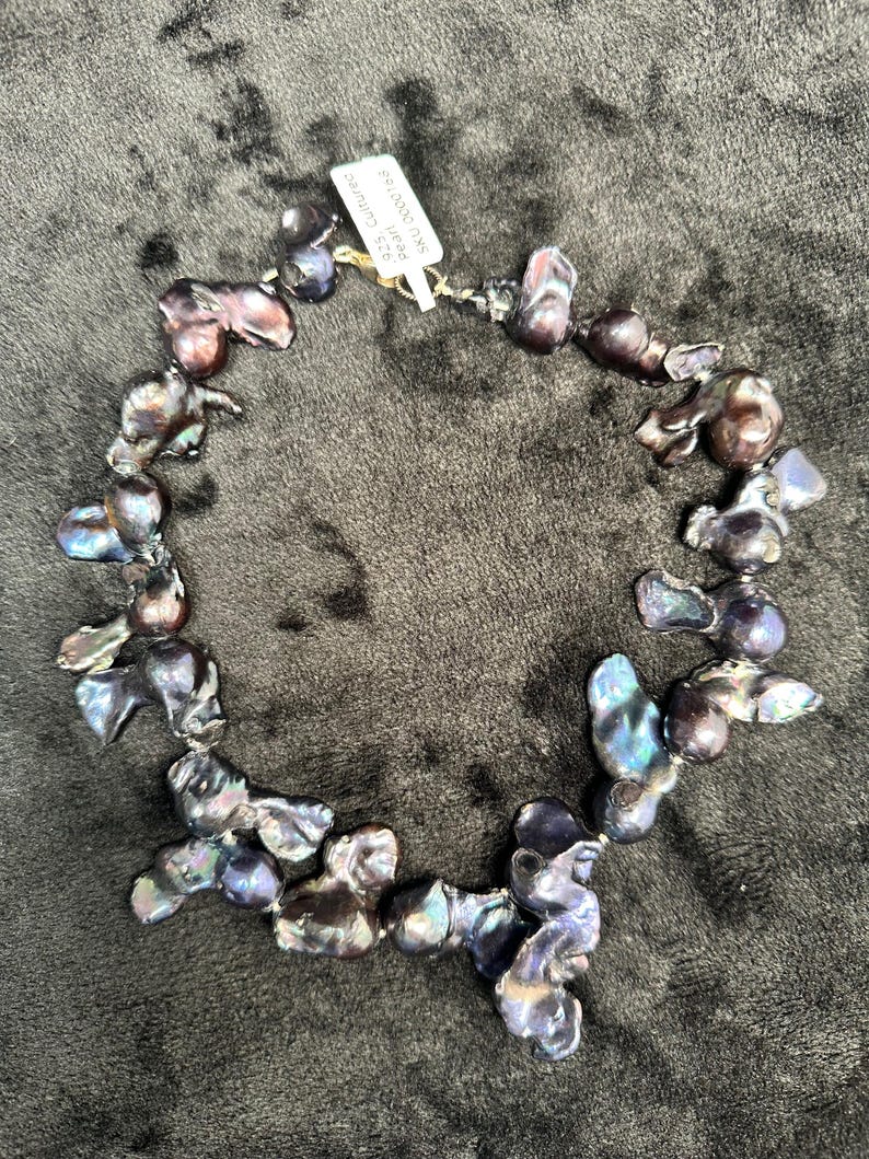 May include: A necklace made of dark iridescent baroque pearls. The pearls are strung on a silver chain and are of varying sizes and shapes. The necklace is displayed on a dark brown surface. The tag attached to the necklace reads "Real Cultured South Sea Pearls 9-10mm".
