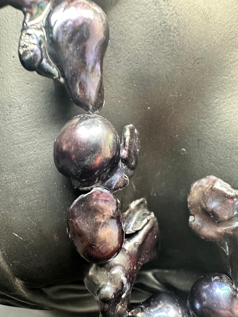 May include: A close-up of a strand of black pearls with iridescent hues. The pearls are various shapes and sizes, some round and some more irregular. The pearls are arranged in a cascading pattern.