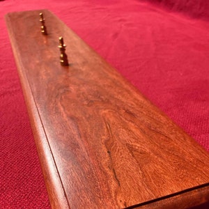 May include: A long, rectangular wooden display with a dark red mahogany finish. The display has five brass pegs along the top edge.