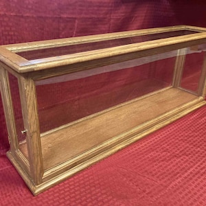 May include: A rectangular display case with a wooden frame and glass panels. The case is made of light-colored wood and has a clear glass top and sides, allowing for full visibility of the contents. The case is designed to showcase collectibles.