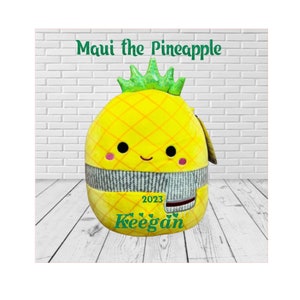 May include: A yellow pineapple-shaped plush toy with a green top and a gray and white scarf. The toy has a smiling face with black eyes and pink cheeks. The text "Maui the Pineapple" is written above the toy, and the text "Keegan 2023" is written below the toy.