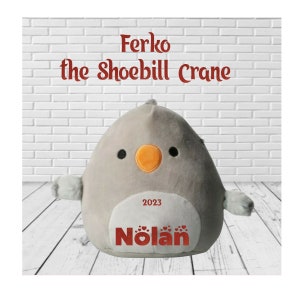May include: A plush gray Shoebill Crane named Ferko with a white belly, orange beak, and black eyes. The text "2023" and the name "Nolan" are printed on the belly. The background is a white brick wall and a wooden floor.