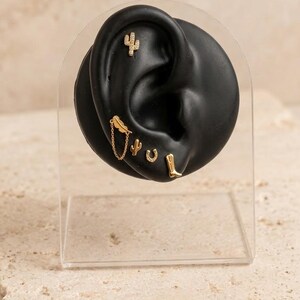 Könnte beinhalten: Ein Set goldfarbener Ohrringe auf einem schwarzen Ohrmodell. Die Kollektion umfasst einen Kaktus-Stecker, einen Feder-Stecker, der durch eine feine Kette mit einem kleinen Kaktus verbunden ist, einen Hufeisen-Stecker und einen Cowboystiefel-Stecker.
