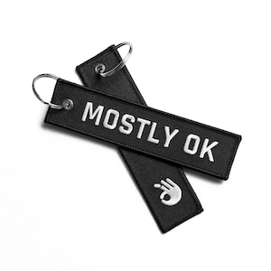 Peut inclure: Deux porte-clés noirs avec des anneaux de clés argentés. Un porte-clés porte le texte "MOSTLY OK" en lettres blanches. L'autre porte-clés a un geste de la main blanc.