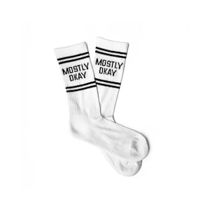 Könnte beinhalten: Ein Paar weiße Socken mit schwarzen Streifen und dem Text "MOSTLY OKAY" auf jeder Socke.