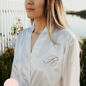 May include: A white satin robe with a personalised embroidered initial "R" in a cursive font. The robe has a sash tie and a V-neck.