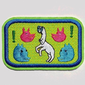 Könnte beinhalten: Gestickter Aufnäher mit grünem Hintergrund und blauem Rand. Der Aufnäher zeigt einen weißen Hund, der über zwei rosa Katzen springt, mit Ausrufezeichen auf beiden Seiten.