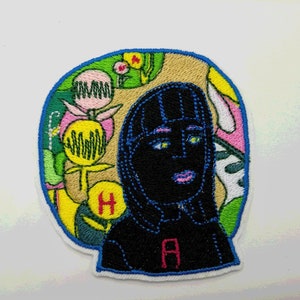 May include: Embroidered patch featuring a black and blue portrait of a person with a colourful floral background. The person has a blue headband and a pink mouth. The letters "HA" are visible on the patch.