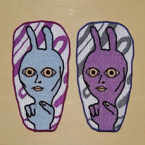 Puede incluir: Dos parches bordados para planchar que representan caras de extraterrestres de dibujos animados con orejas de conejo. Un parche es azul con fondo morado y el otro es morado con fondo gris.