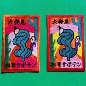 Op de afbeelding: Twee geborduurde patches met een cartoonpotlood, een persoon en de tekst "大発見! 幸せボタン" in het Japans. De patches zijn oranje, rood, geel, groen, blauw en roze.