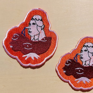 Könnte beinhalten: Zwei gestickte Aufnäher mit orangefarbenem Hintergrund und weißen Umrissen. Jeder Aufnäher zeigt eine Karikatur einer Person mit einer großen Nase und weißen Haaren, die ein braunes Wesen mit großen Augen reitet.