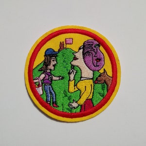 May include: Embroidered patch with a yellow background and a red border. The patch features a cartoon illustration of two people on a green hill with a pink flag. One person is wearing a purple hat and the other is wearing a blue hat.