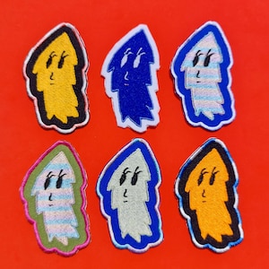 Könnte beinhalten: Sechs gestickte Aufnäher in verschiedenen Farben, darunter Gelb, Blau und Orange, jeweils mit einer stilisierten geisterhaften Figur mit einem lächelnden Gesicht. Die Aufnäher haben einen schwarzen Rand und sind auf rotem Hintergrund angeordnet.