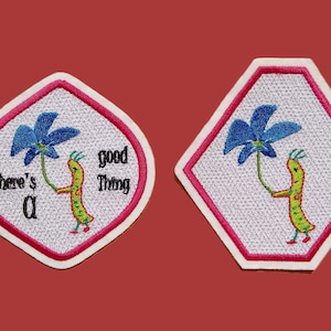 May include: Two embroidered iron-on patches featuring a cartoon worm holding a blue flower. The first patch is shaped like a rounded rectangle with pink trim and the text "There's a good thing". The second patch is shaped like a hexagon with pink trim.