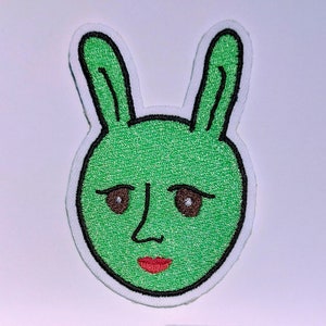 May include: Green embroidered patch of a bunny face with brown eyes and a red mouth.