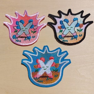 Könnte beinhalten: Drei gestickte Bügelbilder mit einem Comic-Charakter mit einem lächelnden Gesicht und dem Buchstaben "X" in verschiedenen Farben. Die Patches haben die Form einer Flamme mit einem schwarzen Umriss.
