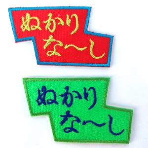 May include: Two embroidered patches with Japanese text. One is red with yellow and blue detailing, the other is green with blue and yellow detailing. The patches have an angular shape and are likely for clothing or accessories.