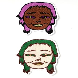 Könnte beinhalten: Zwei gestickte Bügelbilder mit Comic-Gesichtern mit übertriebenen Merkmalen. Das erste Patch hat ein braunes Gesicht mit lila Haaren und das zweite Patch hat ein hellbraunes Gesicht mit grünen Haaren.