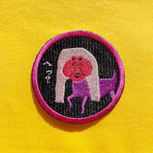 May include: A pink and black embroidered patch with a purple and white dog wearing a pink and white wig. The text "Free" is embroidered on the patch.