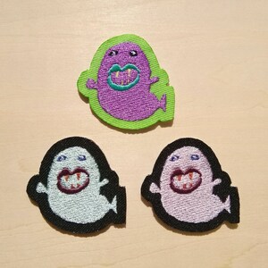 May include: Three embroidered iron-on patches featuring a cartoon ghost with fangs and a toothy grin. The patches are in different colors: purple, light blue, and pink.