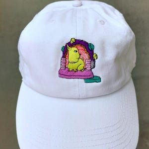 May include: White baseball cap with an embroidered design. The design features a lime green dog-like creature inside a purple and pink structure with yellow accents. The cap has a curved brim and a button on top.