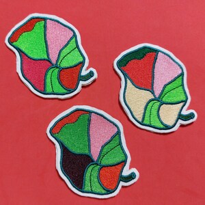 以下が含まれることがあります： 葉の形をした刺繍のアイロン接着パッチが3つ。葉は赤、ピンク、緑、茶色など、さまざまな色のセクションで構成されています。葉は濃い緑色の糸で縁取られています。