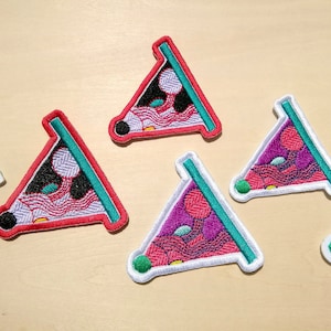 May include: Five embroidered iron-on patches in the shape of a slice of pizza. The patches are in various colors, including pink, purple, black, white, and green. Each patch has a different design, but all feature a wavy pattern and a circle in the center.