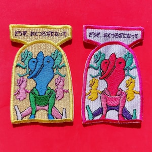Puede incluir: Dos parches bordados con un diseño de dibujos animados. Uno presenta una figura azul con detalles verdes y rosas, el otro una figura roja con detalles amarillos y verdes. Ambos tienen texto japonés en la parte superior y están sobre un fondo rojo.