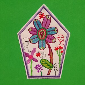 Op de afbeelding: Een witte geborduurde patch met een paarse en blauwe bloem met meerdere ogen, omringd door groene en rode bloemen en drie figuren met ogen.