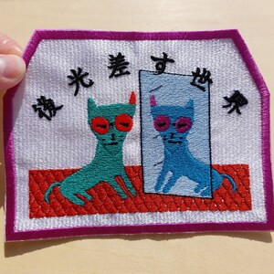 Könnte beinhalten: Gestickter Aufnäher mit zwei blauen Katzen, eine davon schaut in einen Spiegel. Der Text auf dem Aufnäher lautet "夜光美す世物" in schwarzem Faden.