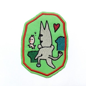Puede incluir: Un parche bordado verde con borde rojo. El parche presenta un gato blanco con un corazón rojo encima, un pequeño pájaro rosa y la palabra japonesa "あ" en negro.