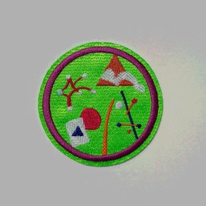 K&ouml;nnte beinhalten: Ein gr&uuml;ner und lilafarbener gestickter Aufn&auml;her mit verschiedenen bunten Formen und Designs, darunter ein roter Kreis, ein wei&szlig;es Dreieck, ein rot-oranges Dreieck und ein wei&szlig;er Stern.