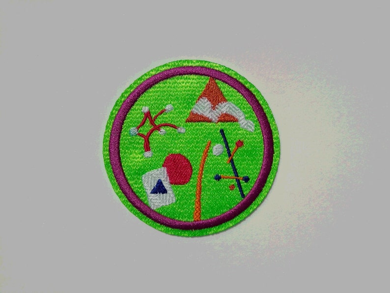 K&ouml;nnte beinhalten: Ein gr&uuml;ner und lilafarbener gestickter Aufn&auml;her mit verschiedenen bunten Formen und Designs, darunter ein roter Kreis, ein wei&szlig;es Dreieck, ein rot-oranges Dreieck und ein wei&szlig;er Stern.