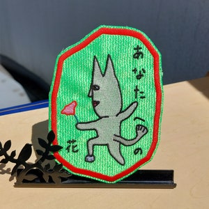 May include: Green embroidered patch with a red border featuring a cartoon creature holding a flower. The creature is gray with a long tail and the text "おやすみ花" is embroidered in black on the patch.