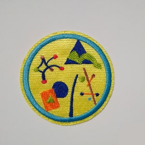 K&ouml;nnte beinhalten: Ein gelber gestickter Aufn&auml;her mit blauem Rand. Der Aufn&auml;her zeigt verschiedene geometrische Formen, darunter ein Dreieck, einen Kreis und ein Rechteck, in verschiedenen Farben, darunter Blau, Gr&uuml;n, Orange und Rot.