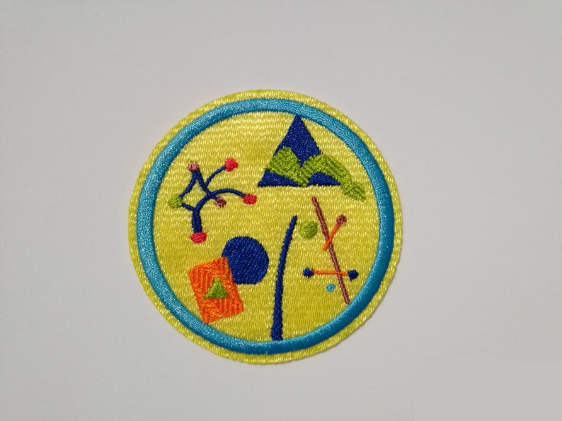 K&ouml;nnte beinhalten: Ein gelber gestickter Aufn&auml;her mit blauem Rand. Der Aufn&auml;her zeigt verschiedene geometrische Formen, darunter ein Dreieck, einen Kreis und ein Rechteck, in verschiedenen Farben, darunter Blau, Gr&uuml;n, Orange und Rot.